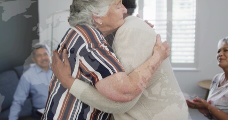 Hugging two senior women wearing striped blouse and knit sweater in living room with sofa, blinds