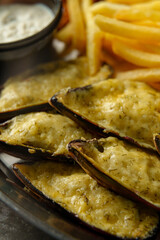 Freshly baked mussels topped with melted cheese sit on a rustic plate, paired with crispy fries and a side of dipping sauce in a warm, inviting seafood restaurant