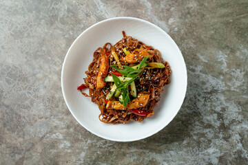 A vibrant bowl holds tender chicken tossed with colorful vegetables and savory noodles. Bright green onions and sesame seeds add a fresh touch, creating a delightful home-cooked meal