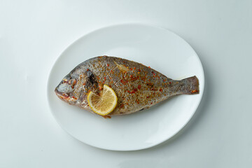 A beautifully cooked fish is presented on a white plate, garnished with a slice of lemon and a sprinkle of spices. This dish offers a tempting presentation perfect for any seafood lover