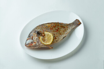 A perfectly grilled fish rests on a clean white plate. The surface is lightly sprinkled with spices, and a bright lemon slice adds a refreshing touch, making it an inviting meal