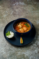 A warm bowl of rich shrimp soup brimming with fresh vegetables and herbs sits on a dark plate, accompanied by a side of fluffy rice and a slice of lime, creating a cozy dining experience