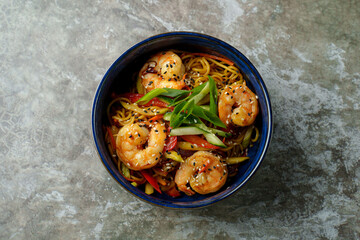 A vibrant bowl filled with tender shrimp and stir-fried vegetables sits atop a textured surface. Noodles are perfectly cooked and garnished with green onions, inviting a delightful dining experience