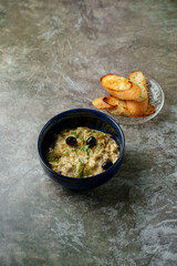 A delicious bowl of creamy, hearty mediterranean dish sits next to crispy toasted bread on a rustic gray table. The olives add a touch of elegance to this delightful meal