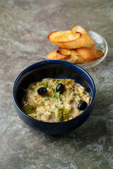 A rich and creamy dip sits in a deep blue bowl, adorned with green herbs and black olives. Crisp toasted slices of bread rest nearby, perfect for dipping