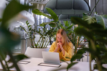 Gen Z freelancer with laptop working in a green coworking space among indoor plants, biophilic design; remote work, focus, productivity, flexible schedule, sustainable work lifestyle.