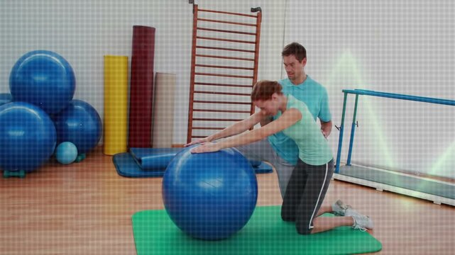 Woman placing hands on blue ball, doing forward reach on green mat, trainer guiding rehab alignment