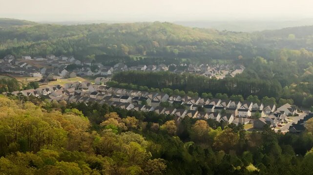 Drone view of a suburban neighborhood surrounded by dense green forests and rolling hills. Homes and streets of residential community nestled on natural landscape