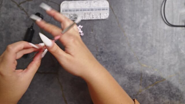 A close-up view of hands applying delicate rhinestone decorations to long, pointed white nails. The detailed process highlights meticulous nail art and beauty care.