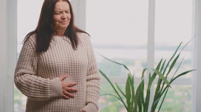 Sick ill elderly woman suffering from menopause period cramps, painful stomach ache on sofa at home room. Sad senior lady holding belly, feeling abdominal menstrual pain. Abdominal pain gastritis