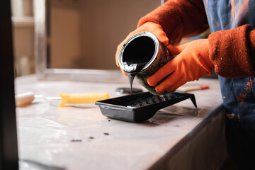 DIY, preparation and supplies concept. Hands in protective gloves pour black paint. Liquid flows from metal can into plastic tray.