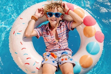 young boy floats in colorful pool on inflatable ring, wearing striped shirt and sunglasses, smiling joyfully, surrounded by clear blue water, joyful summer moment, carefree fun, relaxation