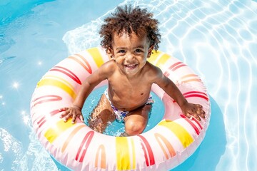 young child with curly hair sits happily on brightly colored striped pool float in clear blue water, joyful family moment, summer fun, carefree child, pool day