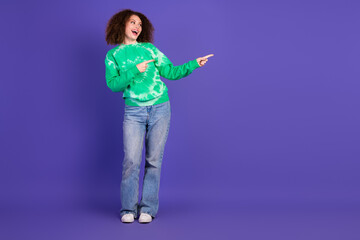 Young woman in a green tie dye sweater jeans smiles and points against a purple background in a casual lifestyle photo