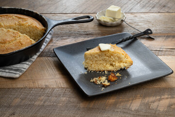 Golden brown, freshly baked, buttermilk cornbread in a cast iron skillet. Slice of bread in a black plate.