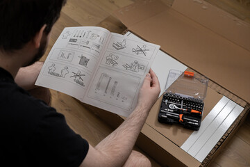 Naklejka premium Person reading a furniture assembly manual with flat-pack components and a toolkit, symbolizing a thoughtful approach to DIY home improvement