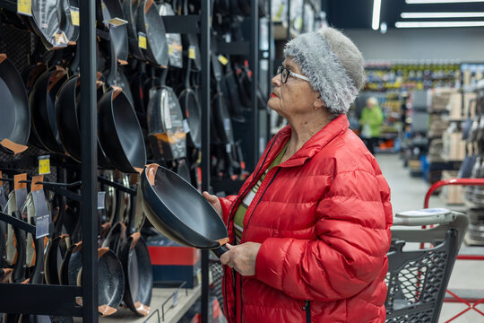 elderly woman shopping independently in a supermarket housewares section, carefully choosing a frying pan from a wide selection of cookware. active senior lifestyle, independent living. 