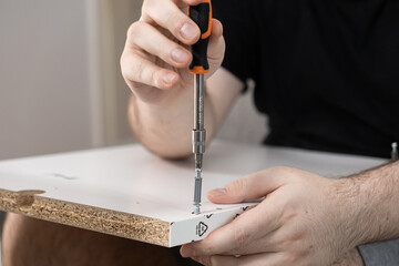 Person assembling white flat-pack furniture board with a screwdriver, fastening metal cam lock...