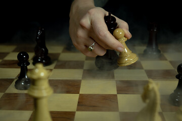 Chess game in progress with a hand moving a piece on a wooden board and smoke effect