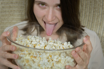 Person holds large bowl of popcorn and makes a funny face while licking their tongue in a casual indoor setting