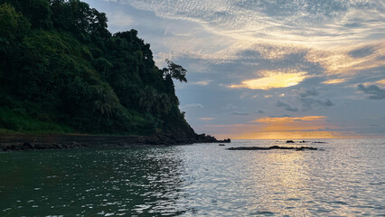 a peaceful coastal scene featuring a lush green cliff beside the ocean. gentle waves hit the rocky shoreline as the sun sets behind scattered clouds