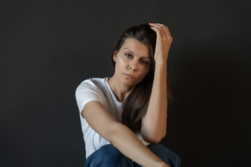 Quiet portrait of woman in casual outfit sitting with arm raised and hand touching hair while gaze turned away creating introspective atmospheric mood captured in soft studio light