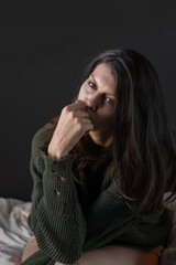 Solitary woman seated on bed wearing green knit sweater looking down in contemplative moody bedroom light capturing intimate quiet moment of introspection and gentle melancholy