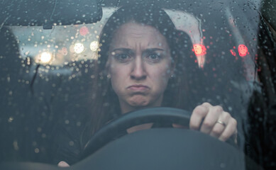 Jeune femme conductrice, l'air triste et contrari&eacute;, au volant de sa voiture sous la pluie avec des lumi&egrave;res de circulation en arri&egrave;re-plan