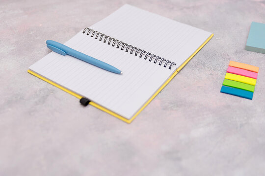 A blank notebook with a blue pen lies on a light gray surface. Colorful sticky notes are arranged nearby, adding a touch of vibrancy to the workspace.
