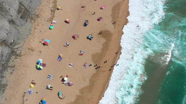 Aerial drone shot of people relaxing on sandy beach surrounded by umbrellas with ocean waves crashing along the coastline on sunny summer day