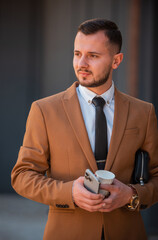 Portrait of a young happy business man in brown suit holding a phone mobile and cup of coffee, near the modern office building.