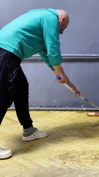 A focused man mops a worn parquet floor, representing cleaning, home improvement, and attention to domestic tasks while wearing a turquoise hoodie and casual Crocs shoes.