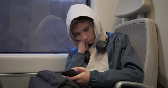 Bored teenage boy with a hood on sitting near a window on a train, leaning on his hand and scrolling on a smartphone screen during a long commute.