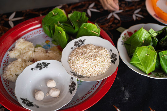 A top-down view of ingredients for a traditional Southeast Asian dessert, possibly Tapai Pulut or Suman Latik. Features glutinous rice, potential yeast balls, and banana leaf wraps on a red and white 