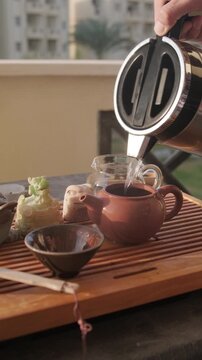 Pouring hot water from metal thermos into small clay teapot set on a wooden tea tray. Warming up a clay teapot. Traditional chinese gongfu tea ceremony at home balcony