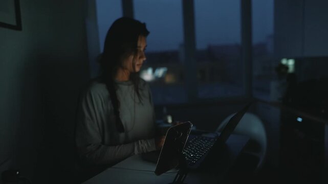 Woman multitasking with phone and laptop under dim city lights