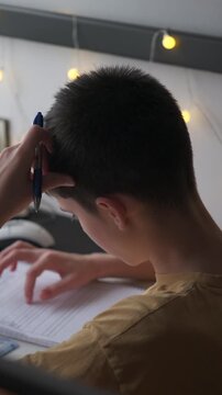 Vertical video of a pensive teenage boy holding a pen to his head while thinking about a difficult homework assignment at a desk in a cozy room.
