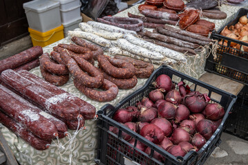A rustic outdoor market stall displays an abundance of traditional Bulgarian cured meats, including various sausages and red onions, presented on a patterned cloth in Bansko, Bulgaria.