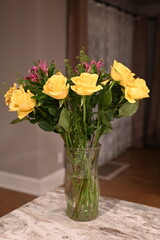 Fototapeta premium Bouquet of fresh yellow roses and pink flowers arranged in a clear glass vase on a table, photographed indoors with warm evening light and a softly blurred home interior.