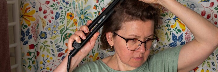 A 50-year-old woman stands in a bathroom with a floral shower curtain. She uses a hair...