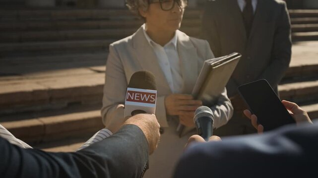 Medium arc shot of middle-aged Caucasian female barrister talking to mass media reporters with microphones while standing outdoors after court hearing, answering questions regarding legal case