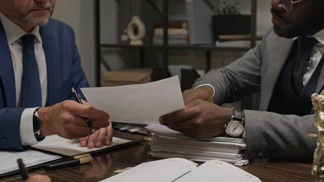 Medium shot of Caucasian and African American male lawyers discussing legal precedent, while reviewing case files during team meeting in office, working on defense arguments