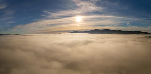 Sun above a sea of fog and clouds with mountain ridge under blue sky, aerial view of foggy mountains