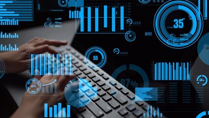 A close-up view of hands typing on a keyboard with overlaying blue digital data visualizations, graphs, and charts depicting various statistics in a modern workspace. Xenic