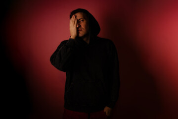 Young man in black hoodie poses with one hand covering his face against a red background in a dimly lit room at evening time