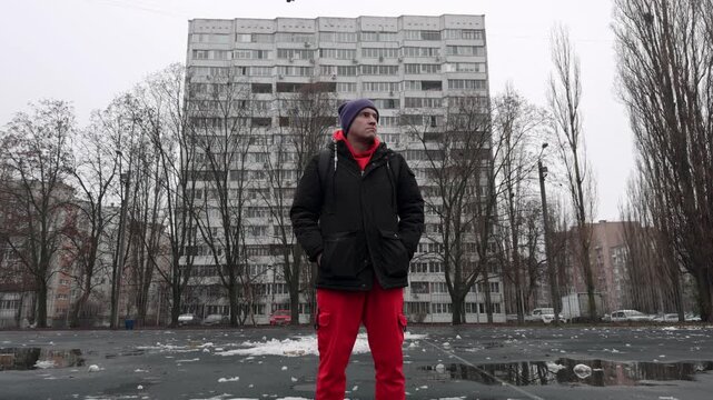 A man is standing in an open space surrounded by tall buildings. The ground is wet, and there is debris scattered around. He looks around while wearing a jacket and beanie.