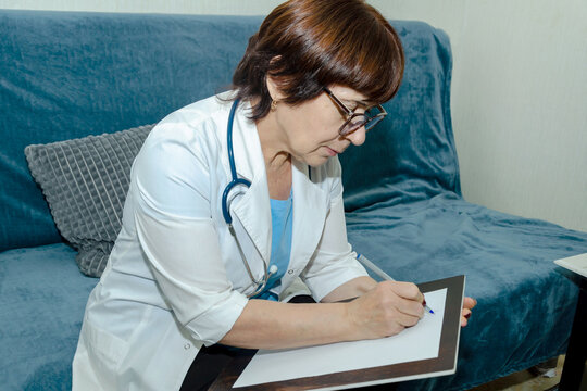 Female doctor in white coat with stethoscope sitting on sofa and writing notes on clipboard after home visit, documenting patient symptoms, treatment plan and follow up recommendations - Powered by Adobe