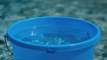 Naklejka premium Blue bucket filled with water, outdoor setting, seasonal activity, cleaning or watering. The object appears to be used for practical household or gardening tasks.
