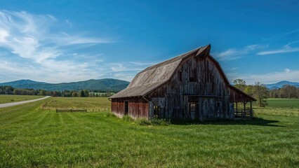 Obraz premium A rustic barn in a lush green field under a bright blue sky with distant mountains.