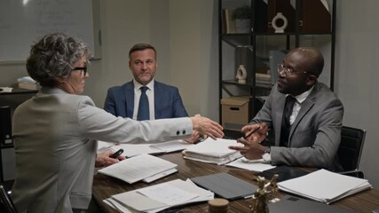 Medium shot of diverse team of corporate lawyers sitting at meeting in office, examining documents, developing defense strategy, agreeing on arguments for court trial and shaking hands